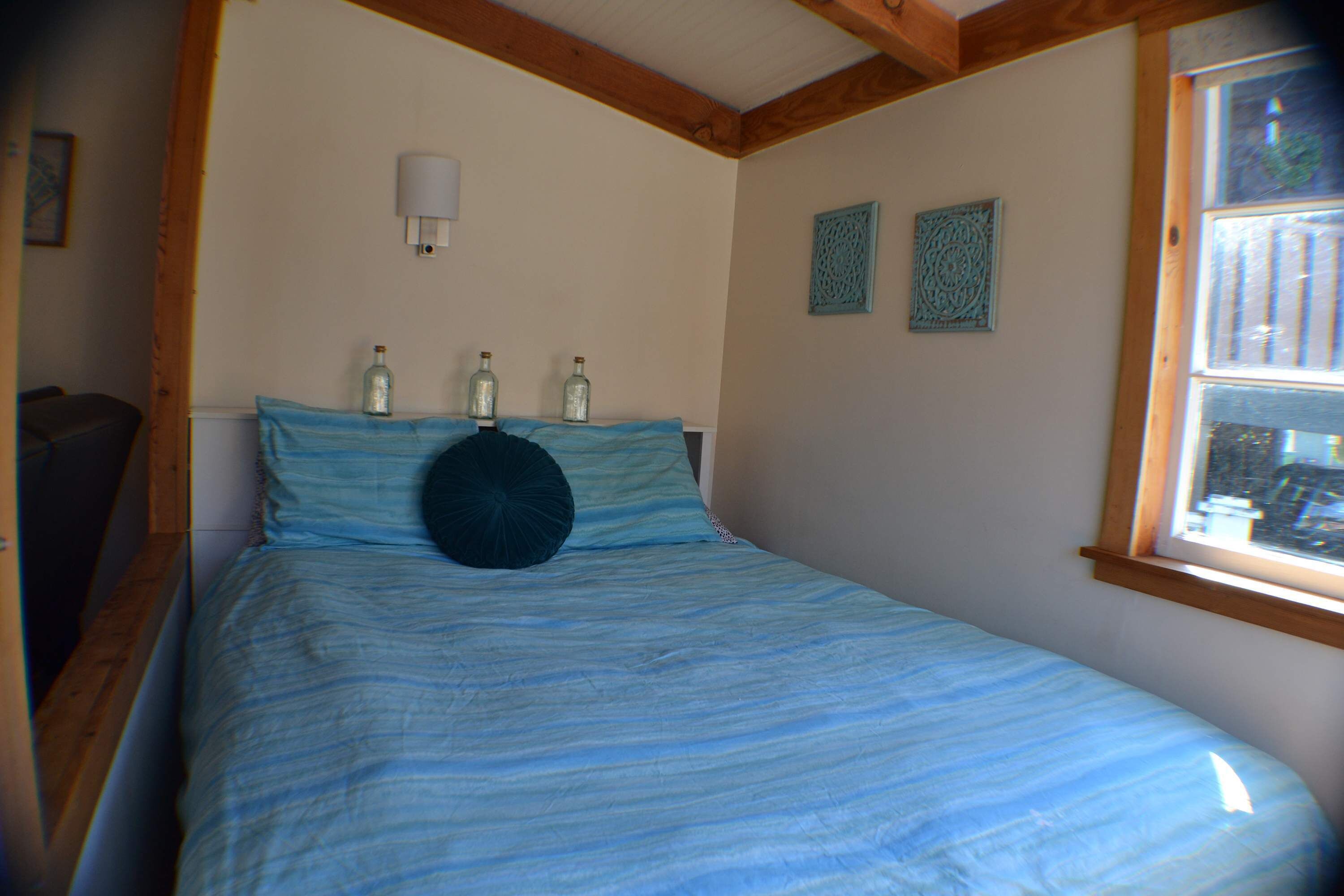 Une chambre à coucher avec un lit, des poutres en bois, une fenêtre et des bouteilles décoratives posées sur la tête de lit.