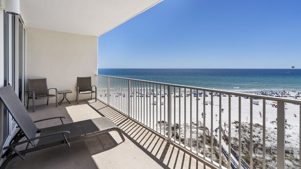 A balcony with two chairs and a table, overlooking a beach with a fence and people. - Lighthouse 502 by Liquid Life (Gulf Shores)