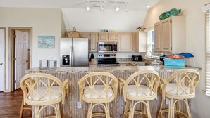 A kitchen with a central island, bar stools, wooden cabinets, and a refrigerator. - Serenity Now: Dog-Friendly Home in Golf Course Community w/ Water Views (Nags Head)