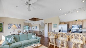 A spacious living room with a sofa, coffee table, and wicker chairs. A kitchen with wooden cabinets and a refrigerator is visible in the background. - Serenity Now: Dog-Friendly Home in Golf Course Community w/ Water Views (Nags Head)