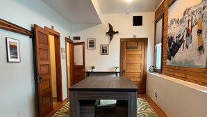 A room with a wooden floor, a dark wooden table, and a green patterned rug. There are two wooden doors, one with a sign above it, and a wall with a framed picture and a mounted bird. - The Office at North Creek All Season Rental (North Creek)