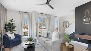A modern living room with a grey sofa, a blue armchair, a round coffee table, and a ceiling fan. There is a large window with curtains, a plant, and a wall-mounted TV.