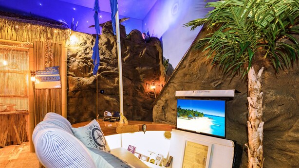 A room with a large screen displaying a beach scene, a bathtub, and a tropical-themed decor including a rock wall and a palm tree. - Shipwrecked-VERY COOL couple's ATL area theme suite! (Carroll County)