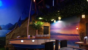 A boat named 'SINKABLE' docked at a beach-themed area with a hot tub and a sunset mural in the background. - Shipwrecked-VERY COOL couple's ATL area theme suite! (Carroll County)