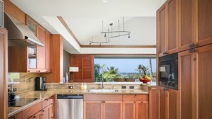 Una cocina moderna con armarios de madera, un lavavajillas de acero inoxidable, un microondas integrado y un amplio ventanal con vista a palmeras y al océano.