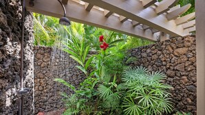 Zona de ducha tropical con pared de piedra, vegetación exuberante y ducha suspendida.