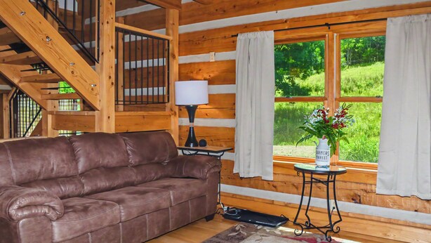 Interior de una cabaña de madera con un sofá de cuero marrón, una lámpara, una mesita, una ventana con cortinas y una escalera.