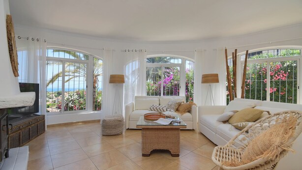 Ein großzügiges Wohnzimmer mit einem weißen Sofa, einem Korbstuhl und einem Glastisch. Große Fenster bieten Blick ins Freie.