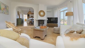 Ein geräumiges Wohnzimmer mit Kamin, weißen Sofas, Korbmöbeln und großen Fenstern.