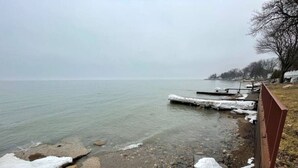 A lakeside view with a rocky shoreline, a wooden fence, and a distant shoreline with buildings. - Krusty Krab! Lakefront Getaway, Walk to Downtown! (Port Sanilac)