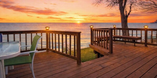 A wooden deck with a table and chairs, overlooking a body of water during sunset.
