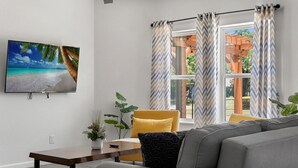 Ein Wohnzimmer mit Sofa, Tisch und Stuhl. Ein Fenster mit Vorhängen, eine Pflanze und ein Bild an der Wand sind ebenfalls vorhanden.