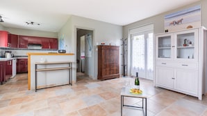 A modern kitchen with red cabinets, a wooden table, and a dining area with a white cabinet and a painting. - La Pêcherie d'Utah - Charmante longère - Proche mer (Audouville-la-Hubert)