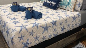Una cama con una colcha estampada con motivos de estrellas, un almohadón con la frase 'elige alegría' y un almohadón con temática de estrellas de mar.