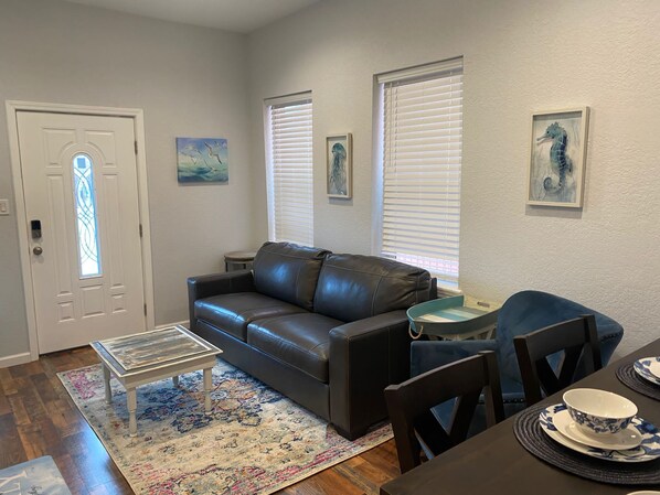 A living room with a leather sofa, a coffee table, a rug, a dining table with chairs, and artwork on the walls. - Cute and Beachy stay for 6, Enjoy walks to beach! (Galveston)