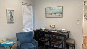 Un comedor con una mesa dispuesta para cuatro, un sillón azul y un cuadro de una escena de playa.