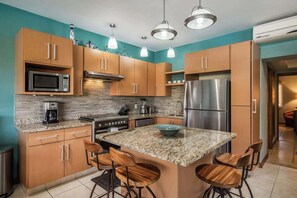 A modern kitchen with wooden cabinets, granite countertops, a central island with bar stools, stainless steel appliances, and pendant lighting.