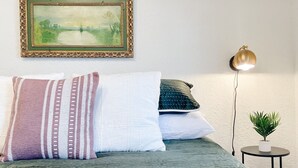 Una cama con colcha verde, varias almohadas, un cuadro enmarcado, una lámpara de pie y una mesita con una planta.