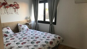 A bedroom with a bed, a nightstand, a lamp, a window with curtains, and wall art of flamingos. - APARTMENT FACING THE SEA, 20 metres from the beach (La Grande-Motte)