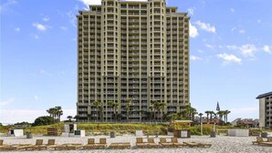 Un hotel moderno con numerosi balconi, una spiaggia sabbiosa in primo piano e palme.