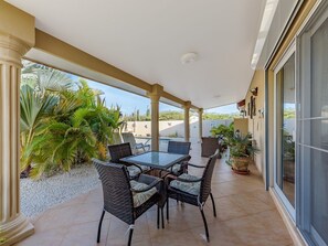 A covered patio with a glass table and wicker chairs, surrounded by potted plants and palm trees.