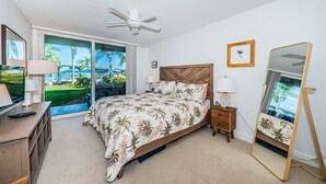 Quarto com cama, televisão, ventoinha no teto, mesa de cabeceira, espelho e vista para uma piscina e palmeiras.