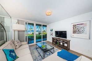 A modern living room with a sofa, a glass coffee table, a flat-screen TV, and a sliding glass door leading to a balcony.