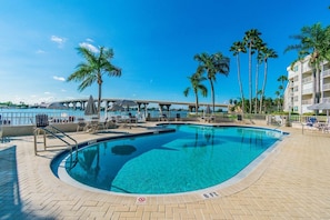 A clear blue pool with a 6 FT depth marker, surrounded by lounge chairs and umbrellas, with a bridge and palm trees in the background.