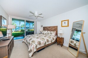 Une chambre d’hôtel équipée d’un lit, d’une télévision, d’un bureau, d’une chaise, d’un ventilateur de plafond, d’une fenêtre donnant sur des palmiers et d’un miroir.