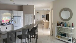 A modern kitchen with white cabinets, a dining area with bar stools, a living room with a sofa, and a bar area with shelves of bottles. - 4-Bedroom Villa with Pool & Jacuzzi, Mangrove View (Port St. Lucie)