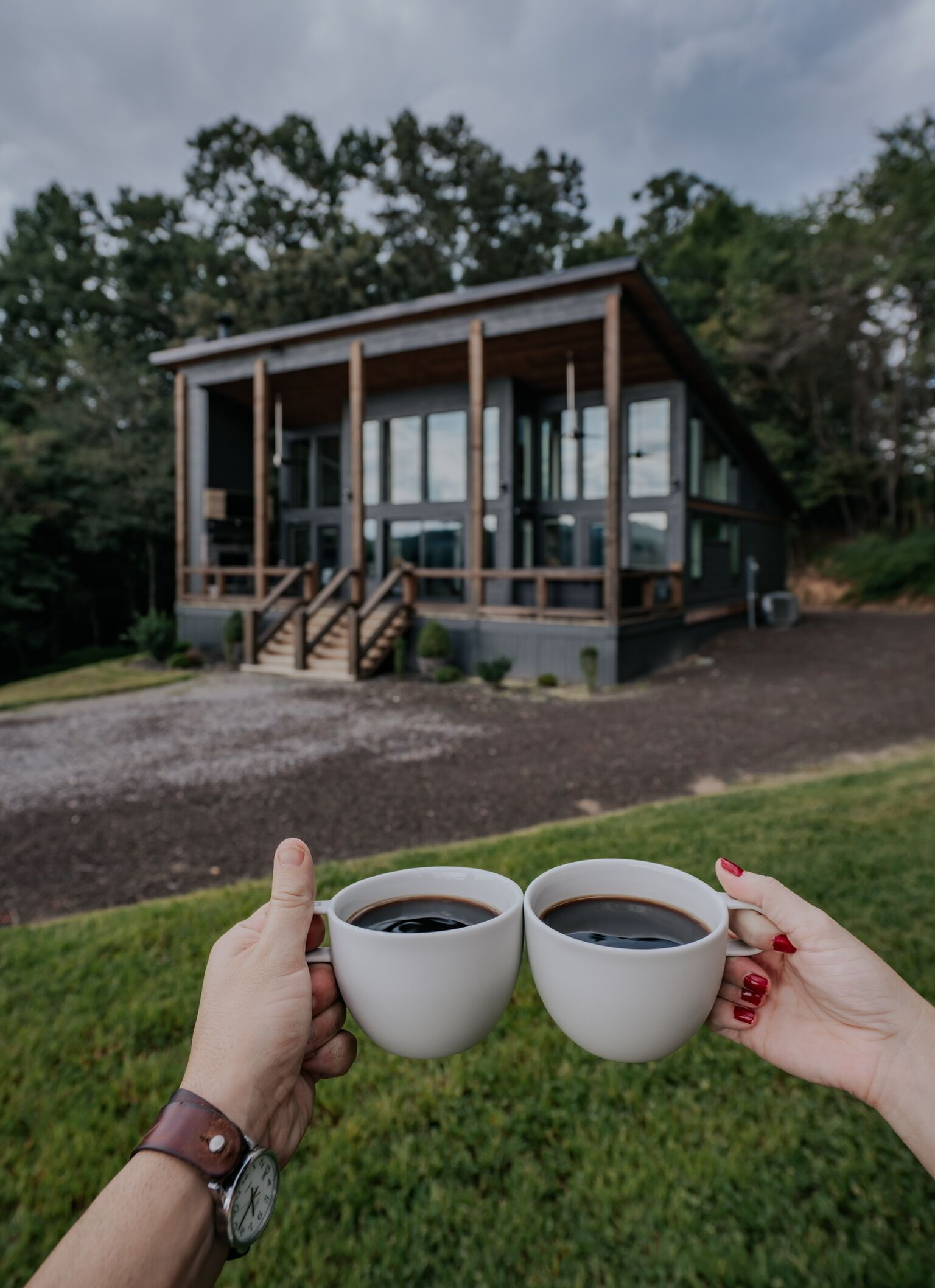 Deux mains tenant deux tasses de café blanc, avec une maison en bois moderne en arrière-plan.