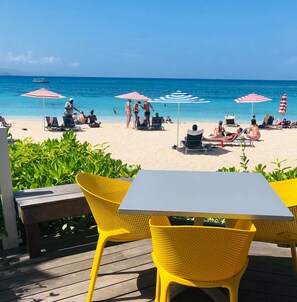 Um alpendre à beira-mar com cadeiras amarelas e uma mesa, com vista para uma praia arenosa com pessoas e guarda-sóis.