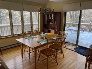 A dining area with a wooden table and chairs, a chandelier, and a view of a snowy deck through large windows. - Amazing family friendly 3+ Bedroom, 3 Full Bath in Quechee  (White River Junction)