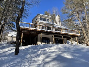 A two-story house with a wooden deck and a covered entryway, surrounded by snow and trees. - Amazing family friendly 3+ Bedroom, 3 Full Bath in Quechee  (White River Junction)