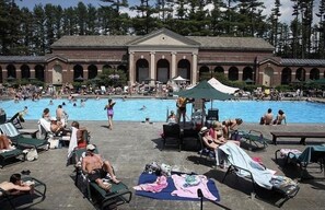 Un área de piscina con sillones de descanso y sombrillas, rodeada por un edificio con ventanales arqueados y una entrada principal.