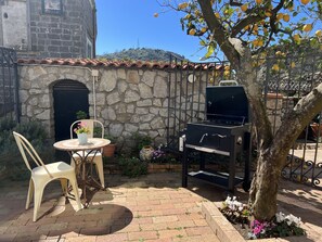 A patio with a stone wall, a grill, a table with a chair, and potted plants.