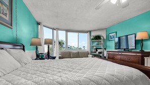 A bedroom with a large bed, a desk, a chair, a bookshelf, and a window with a view of palm trees. - 🌴Beachside Pool~ Onsite Cafe~Studio Top of the Gulf 210 (Panama City)