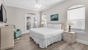 Une chambre avec un grand lit, un ventilateur de plafond, une table de chevet avec une lampe, un fauteuil vert et un tableau représentant une tortue accroché au mur.