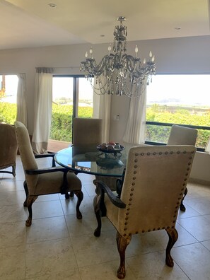 Un espace repas comprenant une table en verre, des chaises rembourrées, un lustre et une vue sur la verdure grâce à de grandes fenêtres.
