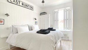 A bedroom with a bed, a sign reading "EAST RUNTON", a radiator, and a window with blinds. - Sea air and starry nights - there's space for two and your four legged friend. (Cromer)