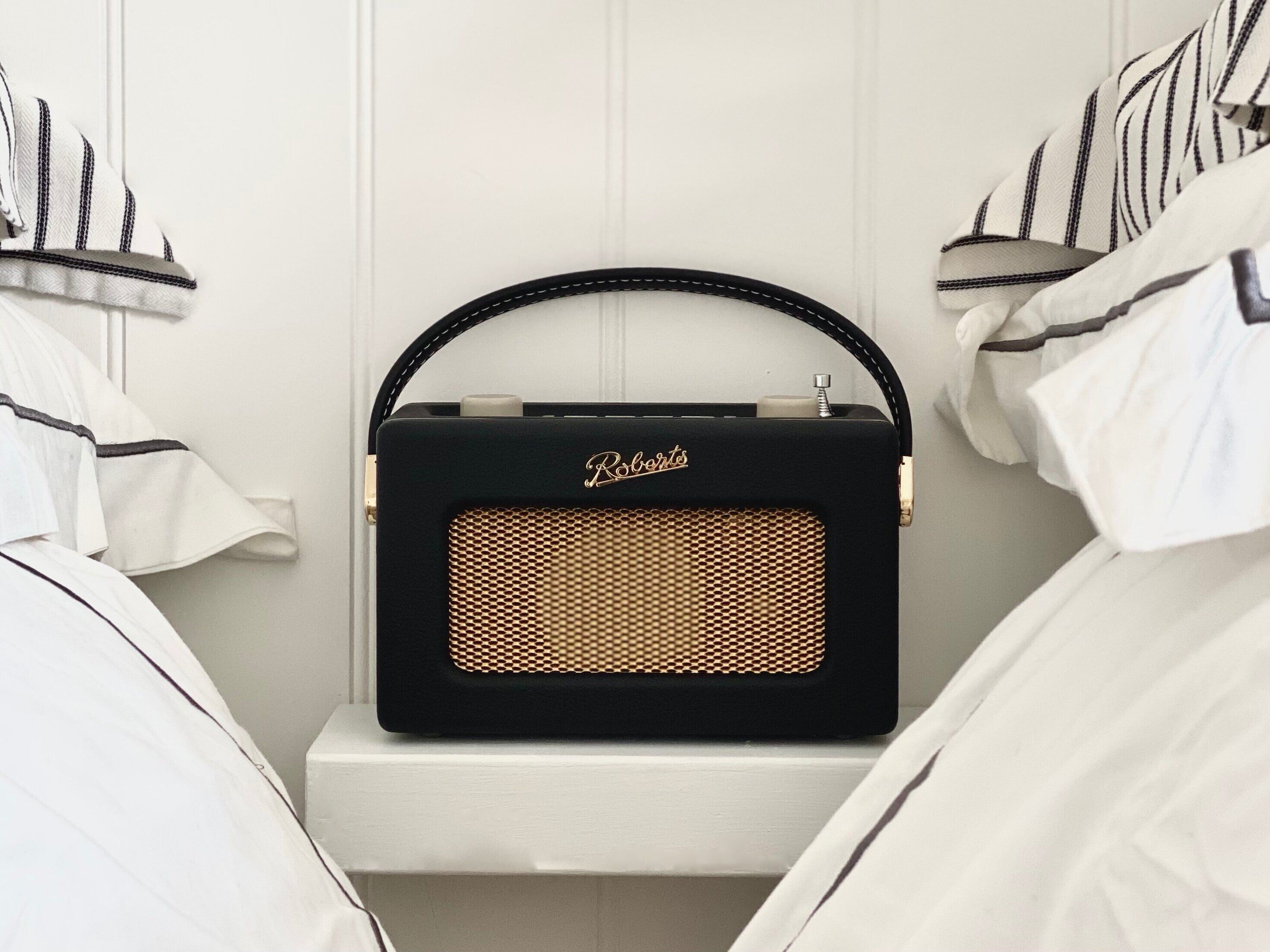Une radio noire portable, dotée d’un haut-parleur à grille dorée, est posée sur une table de chevet blanche dans une pièce où la literie est blanche et les coussins rayés.