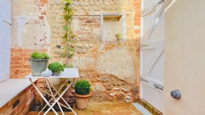A narrow alleyway with a brick wall, a small window, and a white door. There are potted plants on a table and a small garden area. - Sea air and starry nights - there's space for two and your four legged friend. (Cromer)