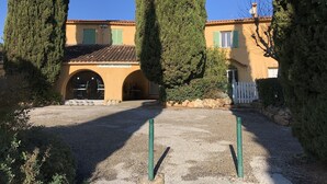 A building with a yellow facade, green shutters, and a tiled roof. There are two large trees in front of the building, and a gravel path leading to the entrance.