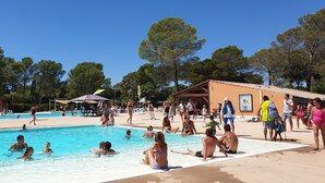 A public swimming pool with people swimming and sunbathing, surrounded by trees and a building with a sign that reads "VOIR LE JOUR".