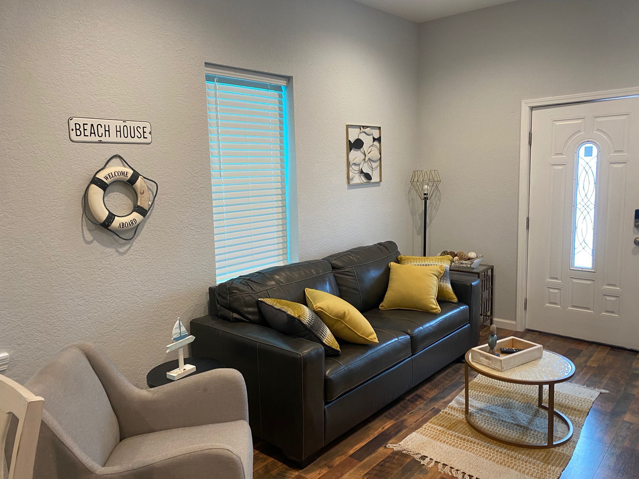 A living room with a black leather sofa, a small round coffee table, and a beige armchair. There is a sign on the wall that reads "BEACH HOUSE" and a life preserver decoration.