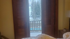 A bedroom with a bed, a dog lying on it, and a view of a snowy outdoor area through open shutters. - Four Seasons Jackson Hole 3-Bedroom March 26 to April 2 Skin-In/out Negotiable (Teton Village)