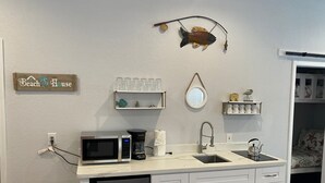A compact kitchen with white cabinets, a microwave, a coffee maker, and a sink. There are decorative items like a fish-shaped ornament and a 'Beach House' sign on the wall. - Great location and Balcony Views! Fun in the Sun (Galveston)
