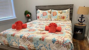 A bed with a floral bedspread, two red towels, a wooden headboard, a nightstand with a lamp, and a small plant. - Great location and Balcony Views! Fun in the Sun (Galveston)