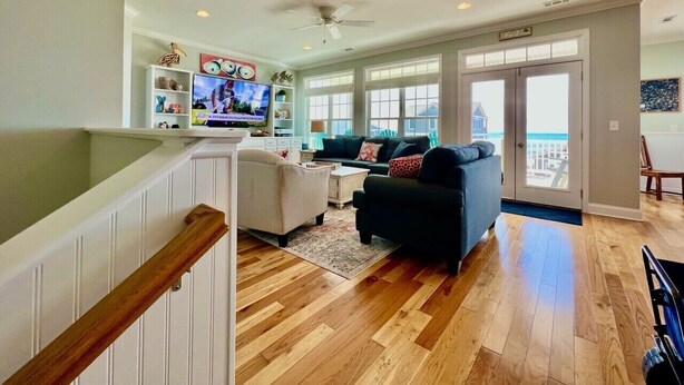 Un salón amplio con piso de madera, un ventilador de techo y un ventanal con vistas al exterior.