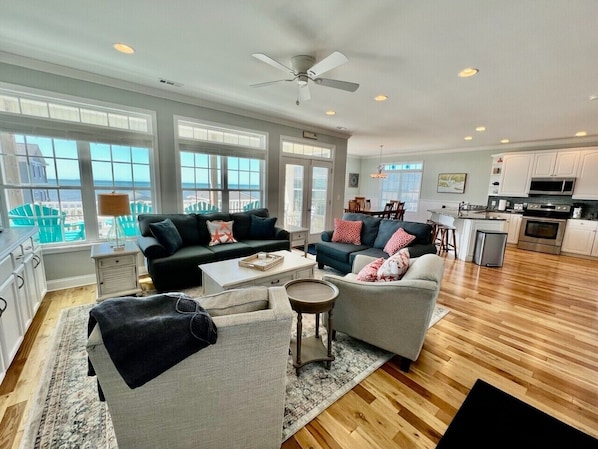 A spacious living room with a large window, a ceiling fan, and a kitchen area visible through the open door.